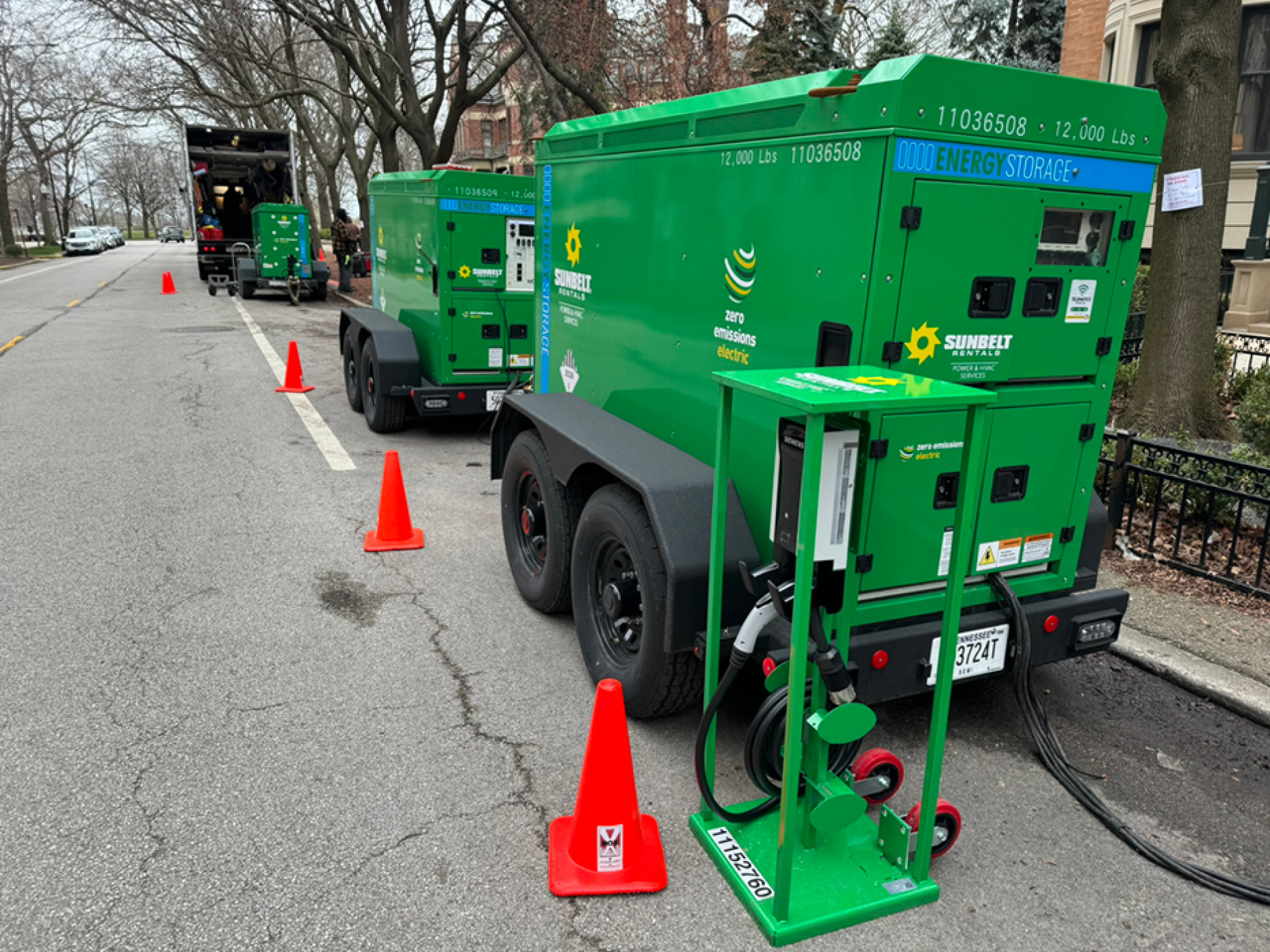 Two Sunbelt Rentals green mobile generators are parked on an urban street, surrounded by orange safety cones.