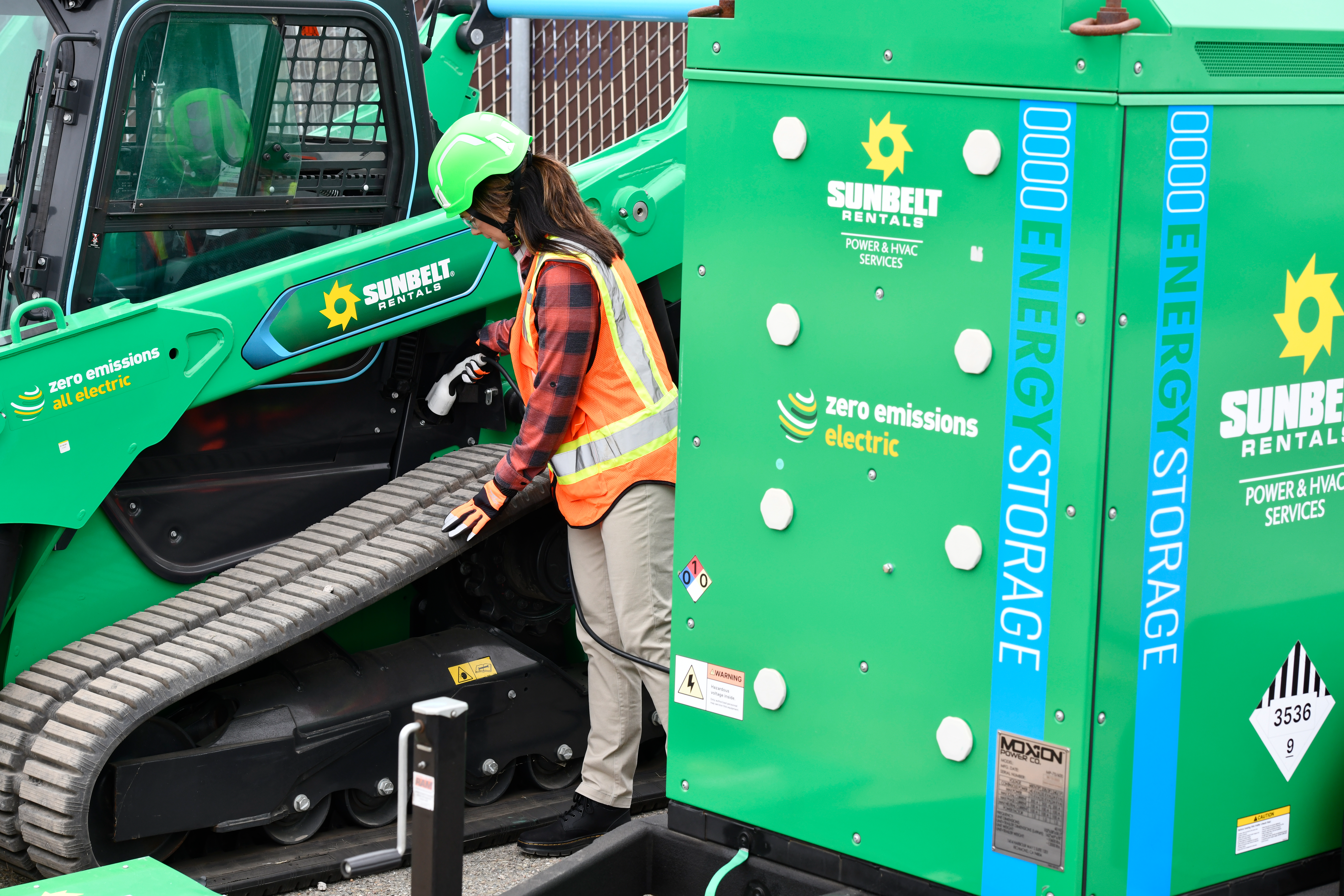 A worker wearing safety gear operates a green Sunbelt Rentals electric machine in an outdoor industrial setting.
