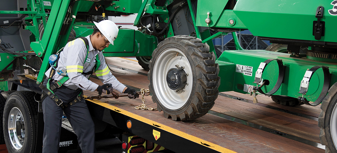 A man, wearing a white safety helmet and green safety gear, safely secures a Sunbelt Rentals manlift to a flatbed truck for an industrial facility maintenance equipment delivery.